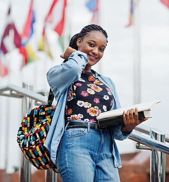 African student female posed with backpack and school items on yard of university, against flags of different countries.